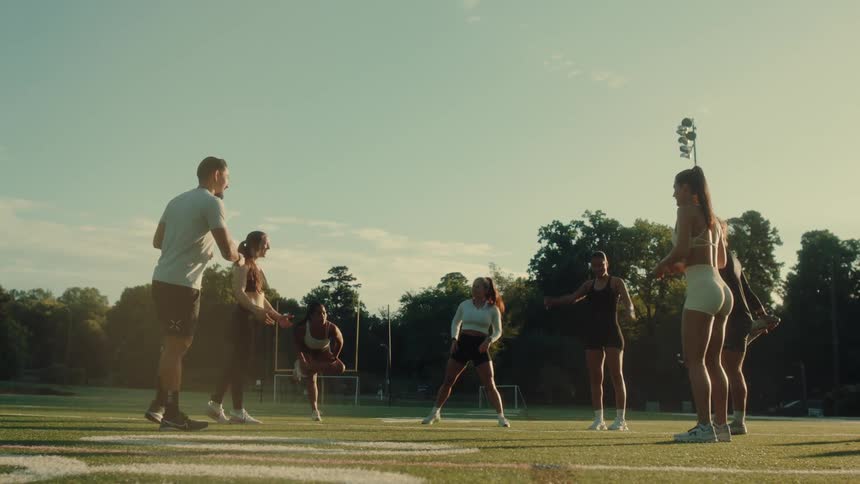 A group of seven young adults exercising on a green athletic field with white lines under a clear sky