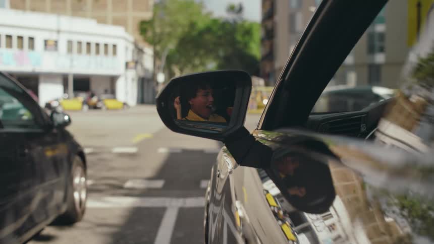 Reflection in car side mirror of a female driver joyfully singing and dancing with her hands to the music while stopped at a stoplight.