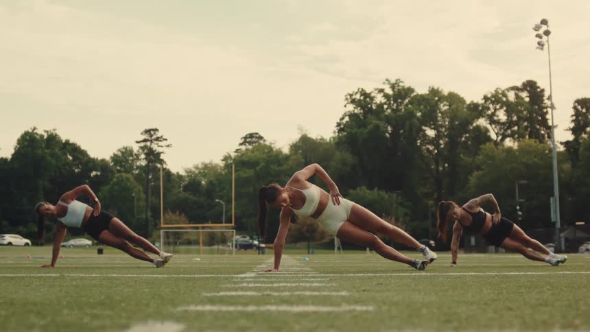 Three women leaning on the grass of an outdoor sports field practicing stretching