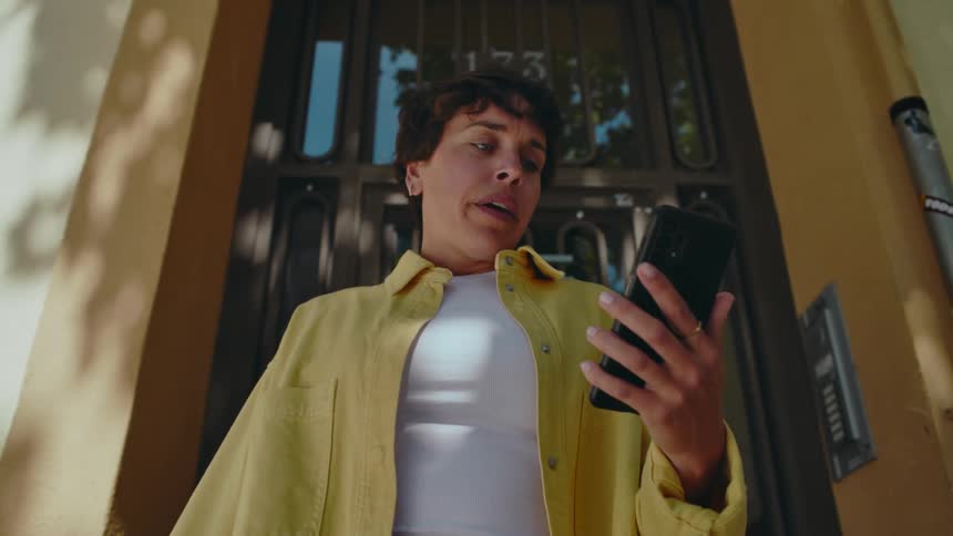 A woman exits her front door looking down at her phone and is yanked away by her dog on a leash that is offscreen.