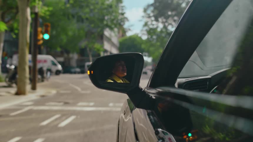 POV looking at the side view mirror as a person drives a vehicle past buildings along tree-lined city streets, parked vehicles, and through an intersection on a sunny summer day