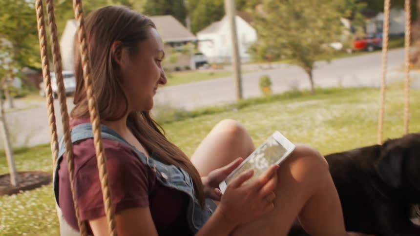Young woman sitting in the backyard of a house interacting with a tablet and with her black dog next to her