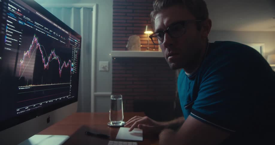 A young man wearing glasses watching a trading chart on a computer monitor in his living room