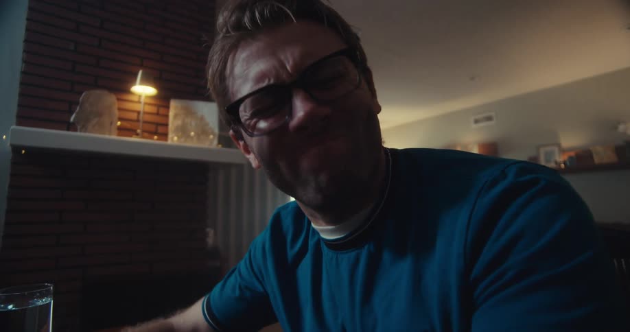 A fairskinned man with reddishblonde hair and glasses wearing headphones and a blue shirt sits at a desk with a computer his expression a mix of frustration and surprise