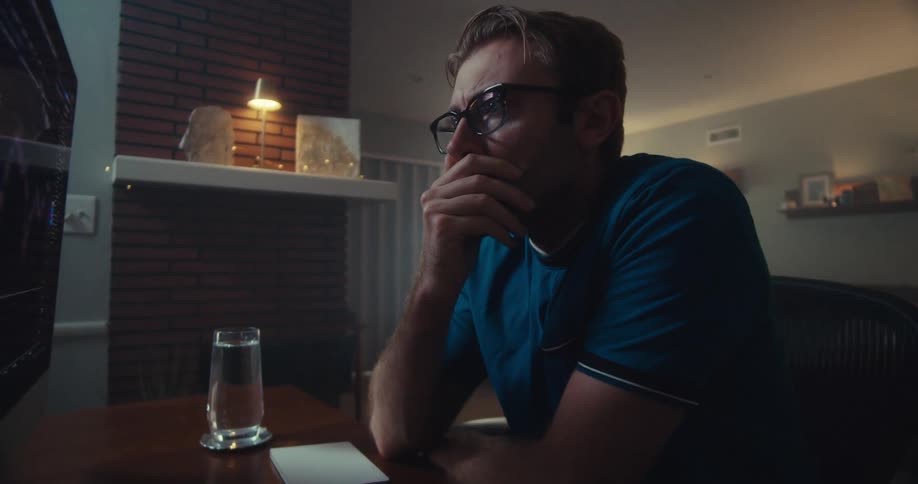 A fairskinned man with light brown hair and glasses looks at a computer screen displaying financial charts in a dimly lit room with a brick fireplace