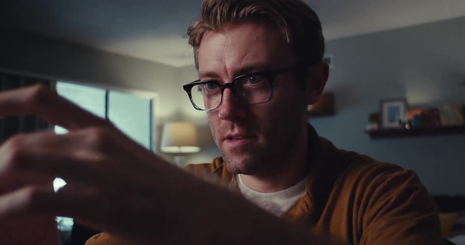 A fairskinned man with light brown hair carefully places a playing card onto a fragile house of cards