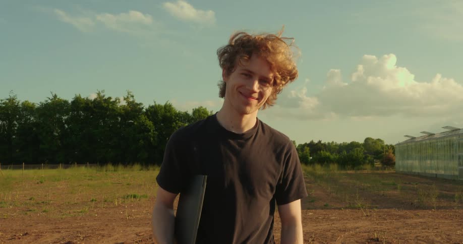 Man stands in a field smiling while holding a computer on a cloudy summer day