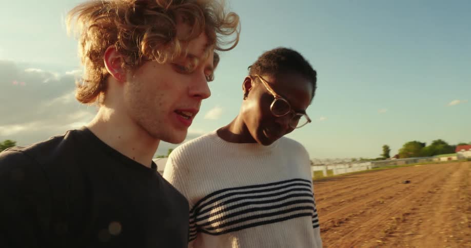 A blond man holding a laptop while talking to a woman who is next to him and they are in an outdoor location with a dirt floor