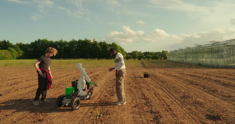 Two people operate an agricultural robot in a field next to a greenhouse on a sunny day