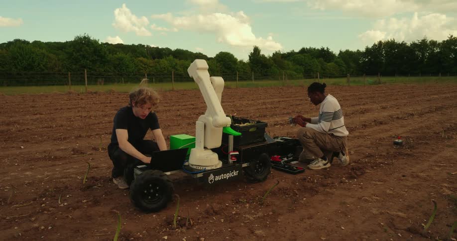Two men are squatting in a field next to a laptop and a robotic piece of farm equipment