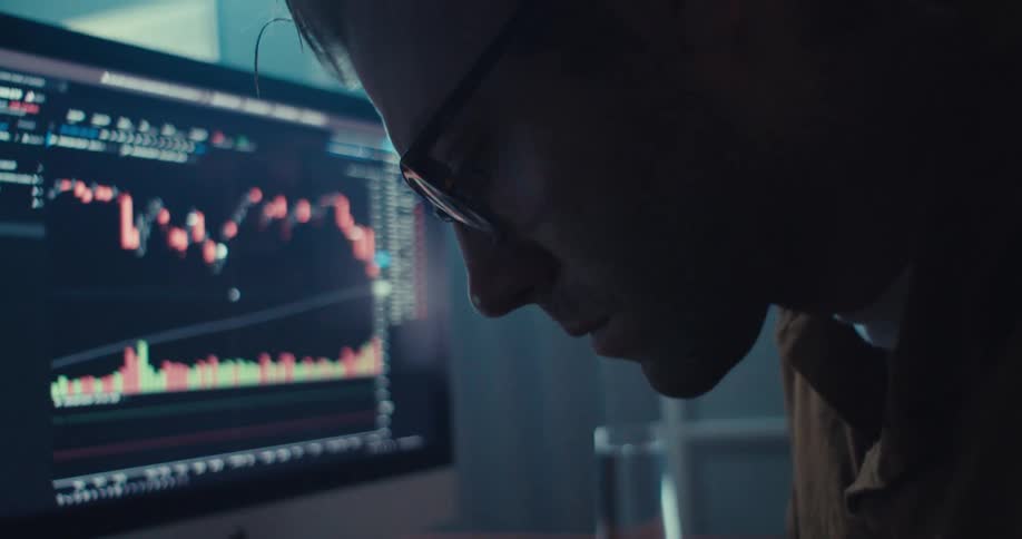 A man wearing glasses intently observes a computer screen displaying financial charts in a dimly lit room