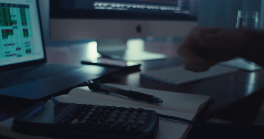 A man looking at a notebook tossing a pen onto it then turning to a monitor displaying binary options