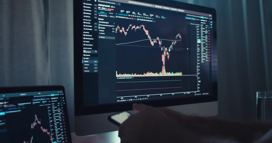 A persons hand holds a smartphone displaying financial data in front of two computer screens also showing stock charts