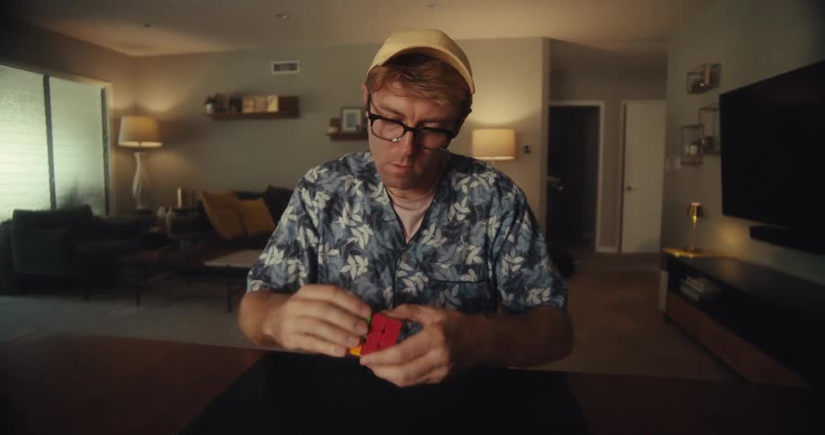 A man wearing glasses and a cap sits at a table solving and looking down at a rubiks cube