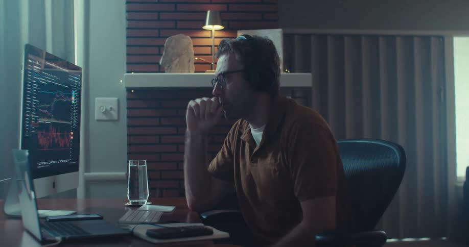 A man sitting at a desk looking at a computer screen analyzing a binary options chart and speaking