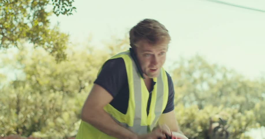 A person wearing a safety vest stands outdoors holds a phone to their ear finishes the call and puts on a red hard hat