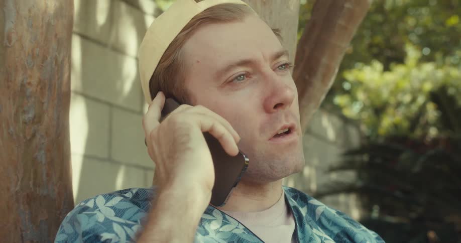 A man relaxes under a tree in a backyard while having a cheerful phone conversation