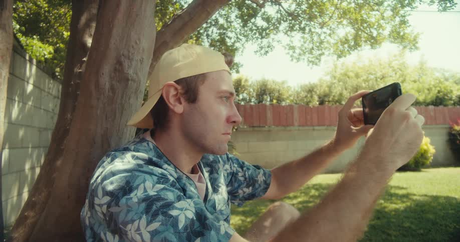 A young man wearing a hat sits in his yard beside a tree on a sunny day using his smartphone to take video footage of his yard