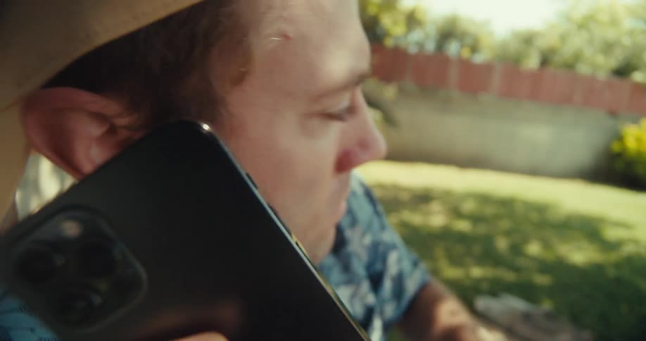 A man answers his cell phone while sitting by a tree