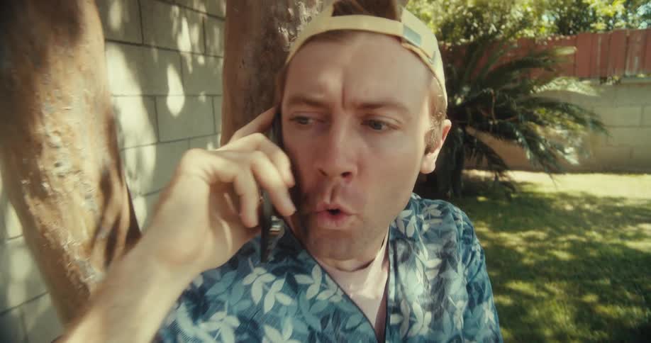 A man sitting under a tree outdoors talks on the phone smiling before appearing slightly worried