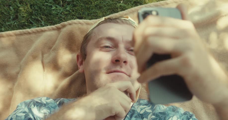 Man types on his mobile phone while laying on his back outdoor at midday