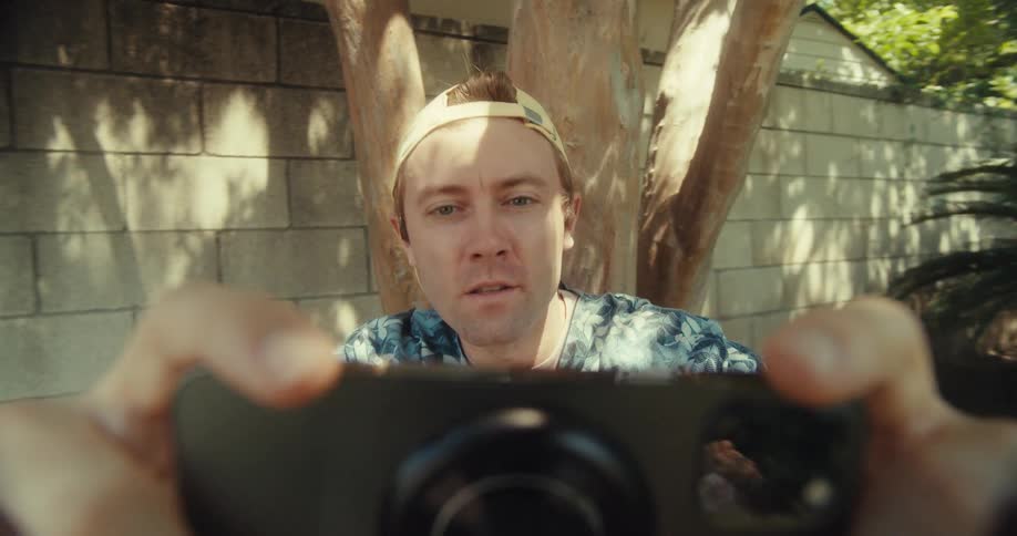A man wearing a baseball cap holds a camera up to take a picture outdoors