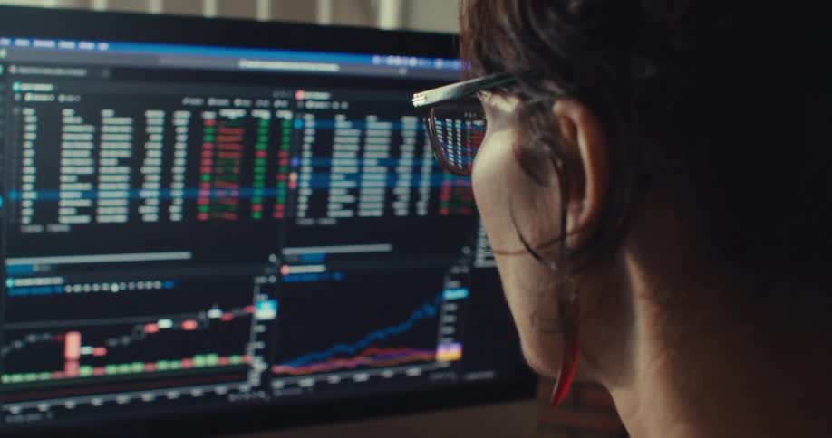 A woman closely monitors stock market data and analytics on a large computer screen