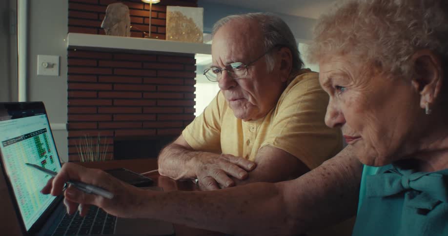 An elderly couple studies information on a laptop screen while sitting in a living room