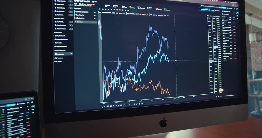 Closeup of a laptop and computer screen displaying fluctuating stock charts in a trading setup