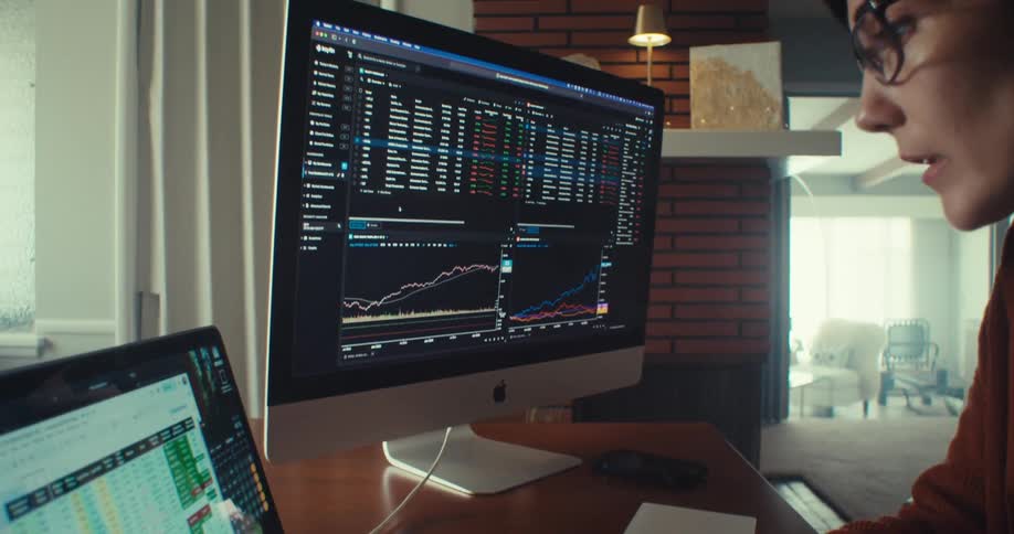 A woman is sitting at a desk looking at two monitors with graphs on them