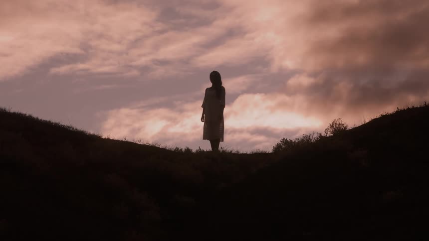 A woman stands alone silhouetted on a hilltop at sunset with dramatic clouds behind her