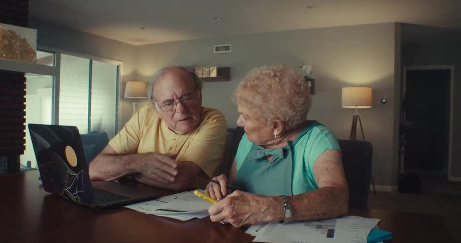 An elderly man and woman are sitting at a table going through papers and there is a laptop in front of them