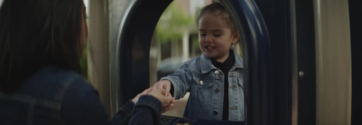 A young child with curly hair and a bow plays inside a blue structure at a playground observed by an adult