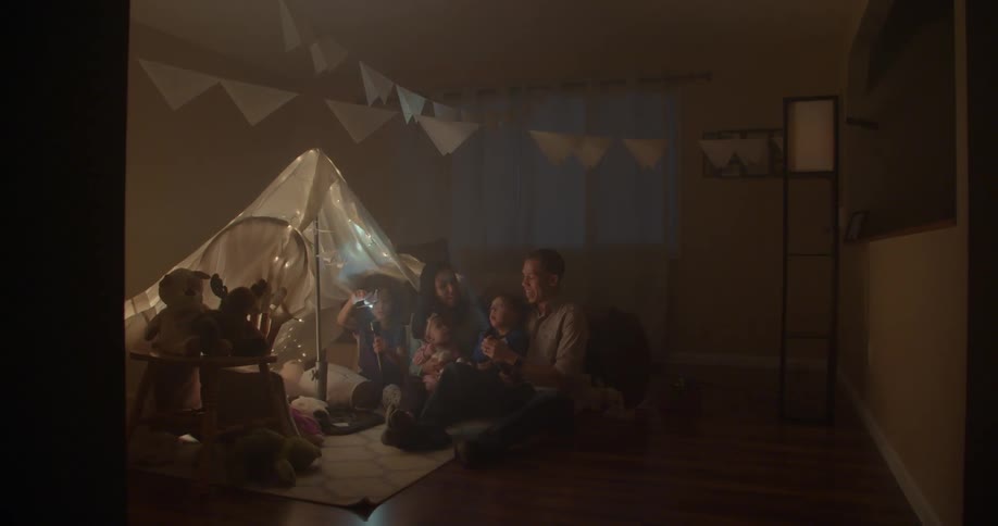A family sits on the floor near a blanket tent with flashlights