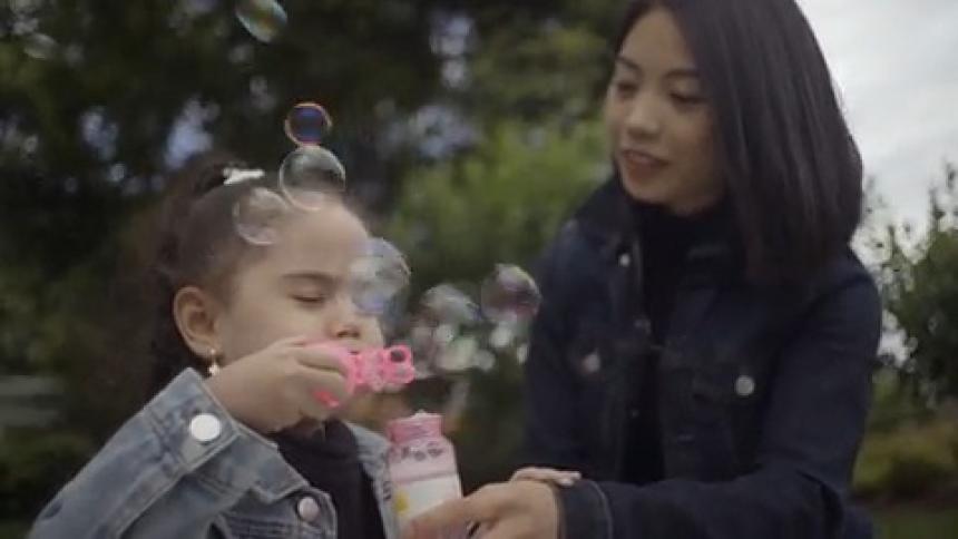A little girl blows bubbles with her mother among some large trees