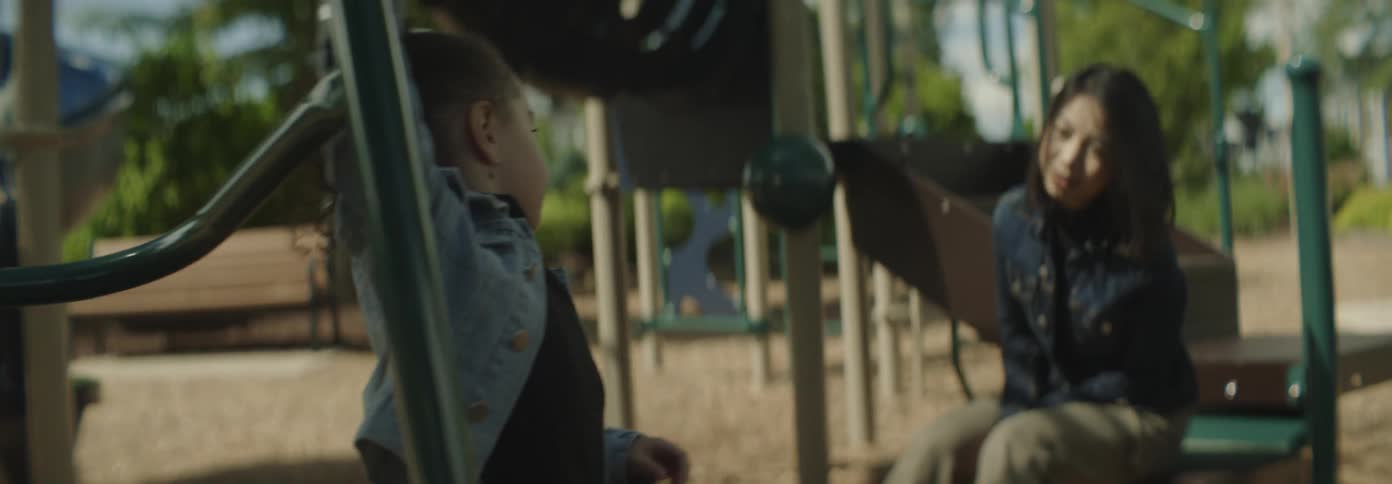 A little girl runs up to her mother who is sitting in a playground by a slide