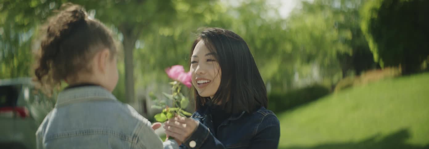 An asian mother squats and waits for her daughter at midday smiling brightly as she hugs her child who brings her a flower