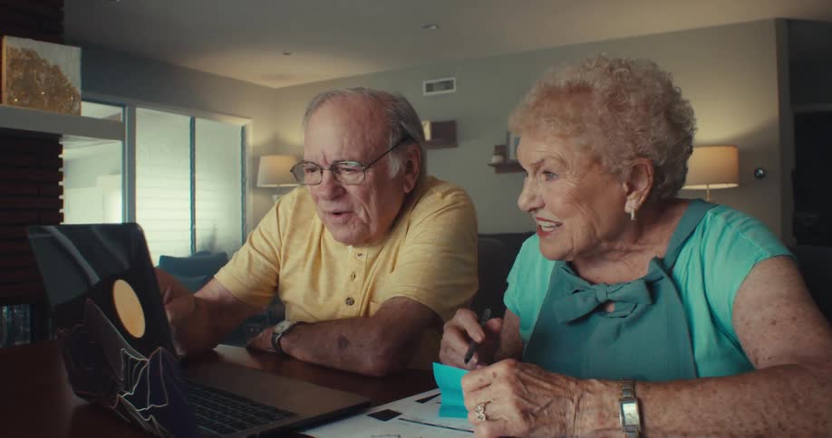 An elderly woman sits beside her husband indoors as they watch a computer screen happily discussing while she writes down notes on a piece of paper