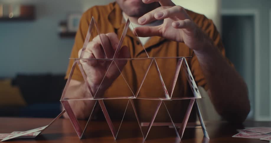 A man wearing glasses carefully stacks a deck of cards but when the cards stumble he gets angry and hits the desk