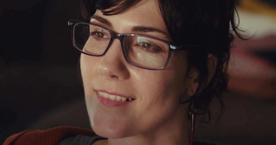 A woman with short dark hair and glasses smiles gently as she looks off screen with light reflections visible in her lenses