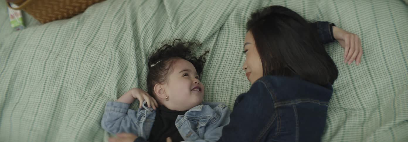 A woman with dark hair and a young girl with curly hair laugh and look at each other while lying on a textured green blanket