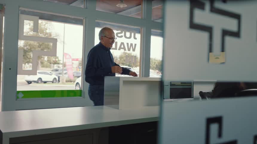 An older man wearing glasses stands at a counter talking to someone seated behind a computer