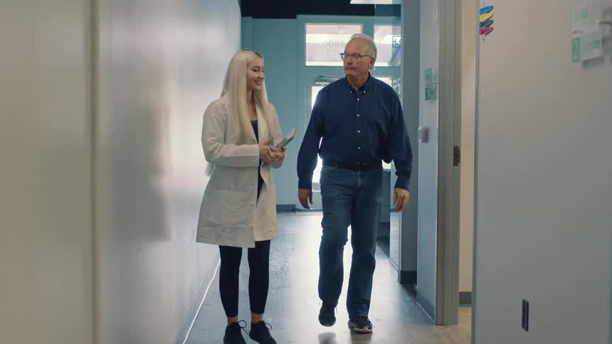 A female healthcare worker and an older man walk and talk in a hallway