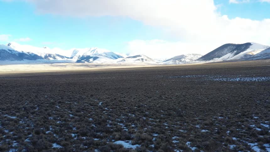 Two horses gallop across a sparse snowdusted landscape with snowcapped mountains rising in the distance beneath a partly cloudy sky