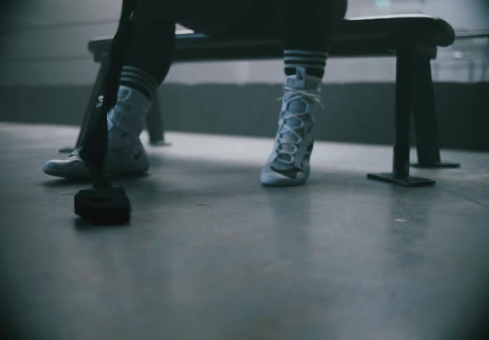 Black boxing hand wraps are dragged across the gym floor as a person pulls them with an overlit boxing ring visible in the background