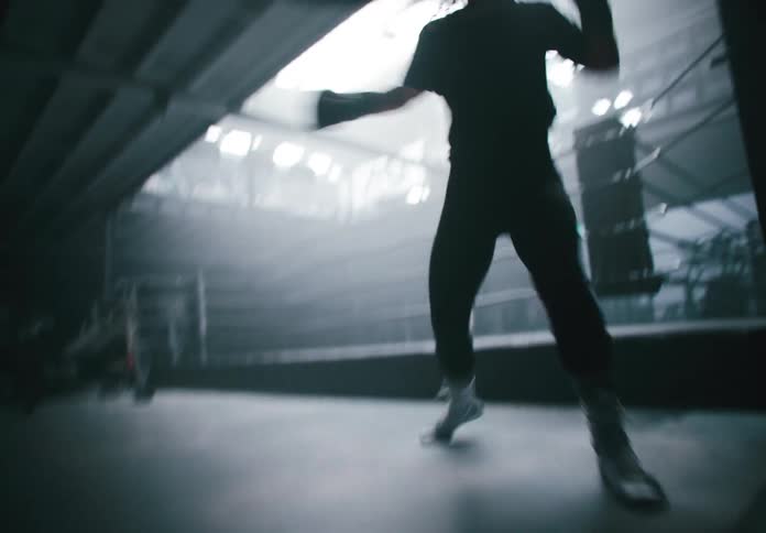 A person in boxing gloves and boots moves around a boxing ring shown mostly in silhouette against the light from above