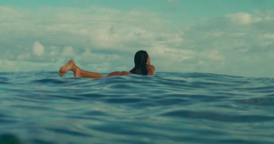 Female surfer lying on her surfboard uses her hands to paddle it out to sea on a cloudy summer day
