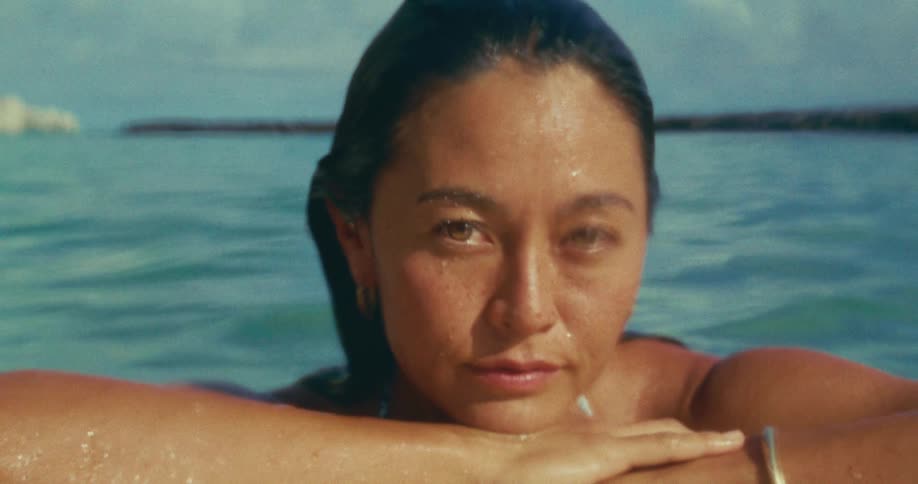 A woman rests her arms on a surfboard while looking directly into the camera