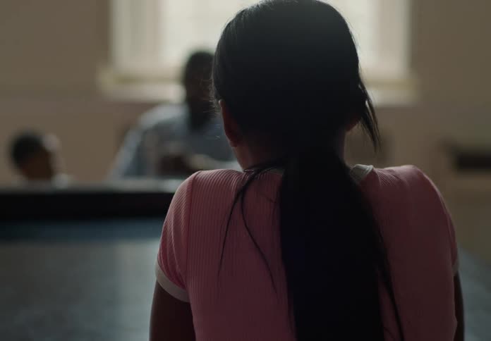 A child with long dark hair in a ponytail and a pink shirt reclines on a piano facing away from the camera with blurry figures seated in the background