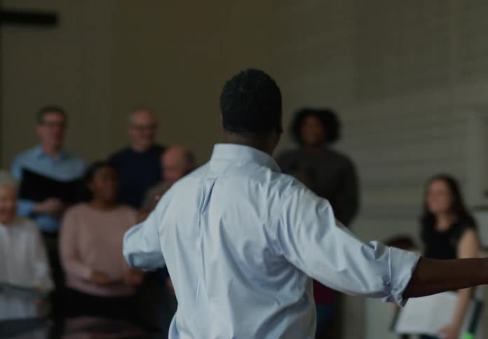 A choir director enters a church and stands facing a group of choir singers with a cross visible in the background